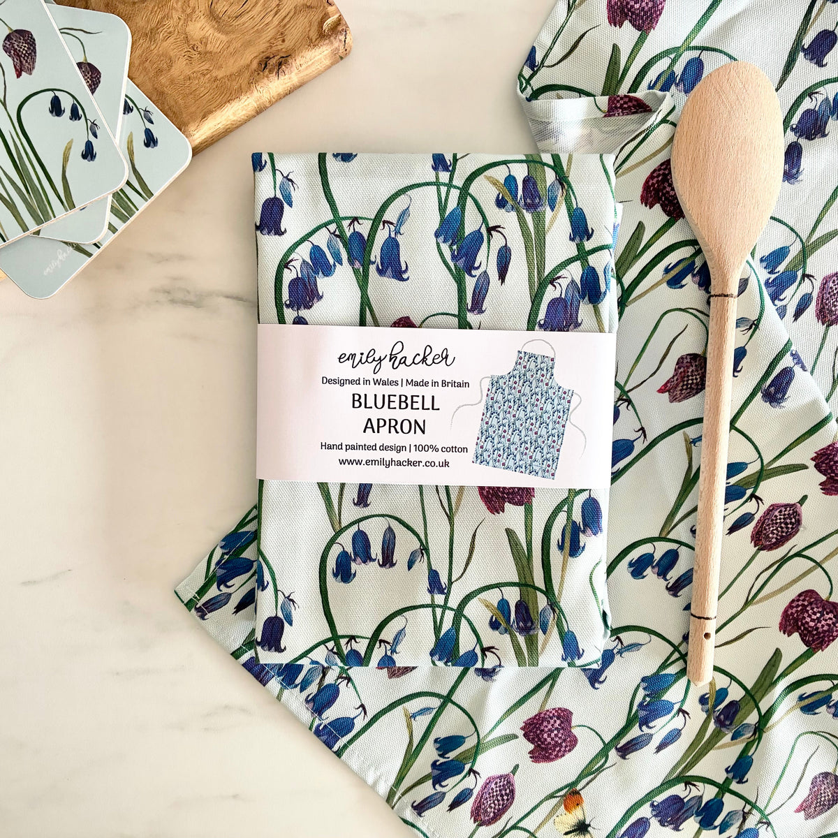 An apron with a bluebell and snake head fritillary flower pattern laid on a light marble surface next to a wooden spoon, matching coasters, and a wooden cutting board. The package label reads 'emily hacker' and 'BLUEBELL APRON'.