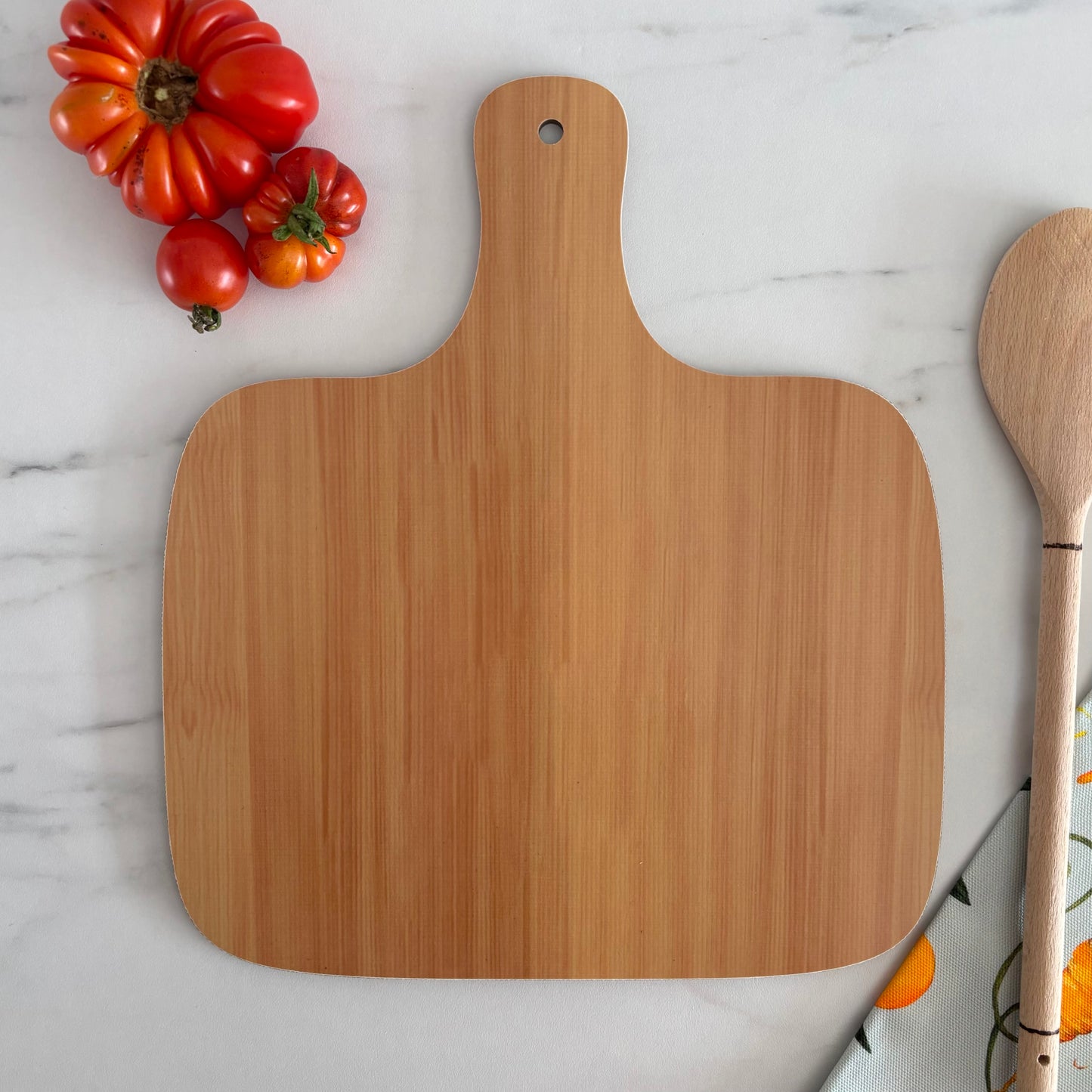 Woodgrain chopping board back photographed on a marble background next to wooden spoon and tomatoes