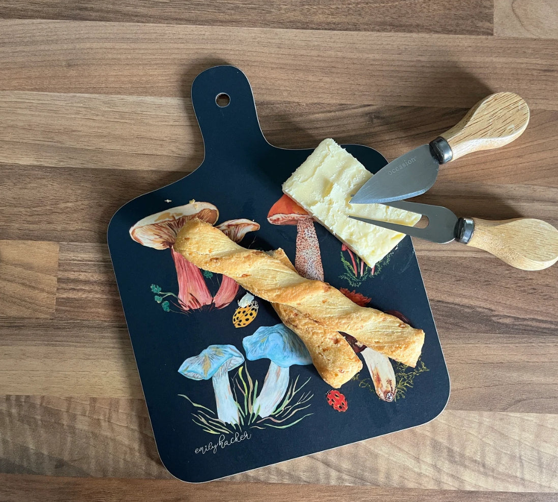 A small dark blue chopping board with hand painted illustrations of mushrooms and a ladybird, accompanied by a cheese knife and fork.