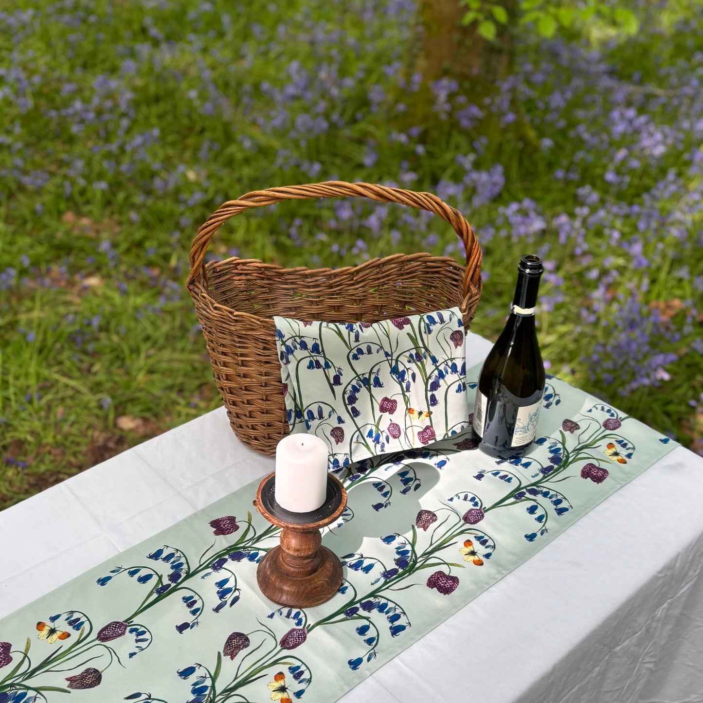 Outdoor setting with a floral bluebell cotton table runner, bottle, candle, and basket in a natural setting.