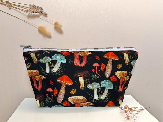 A black washbag with hand painted illustrations of various mushrooms in colors red, orange, and gray, and ladybirds on it. The washbag has a zipper and is placed on a white surface with some dried plants at the top.
