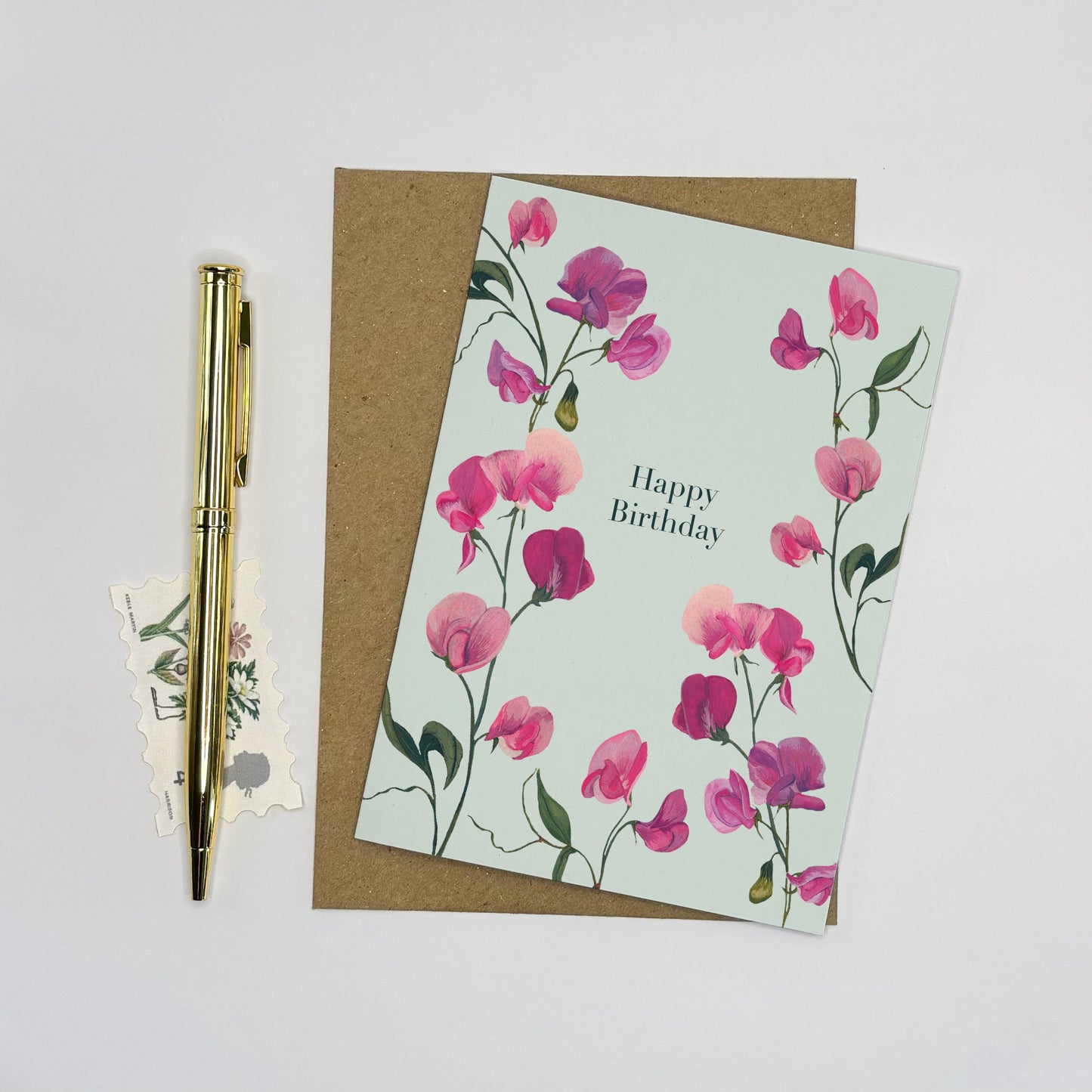 Floral sweet pea happy birthday card with pink flowers on a light green background, placed on a white surface with a pen and additional cards.