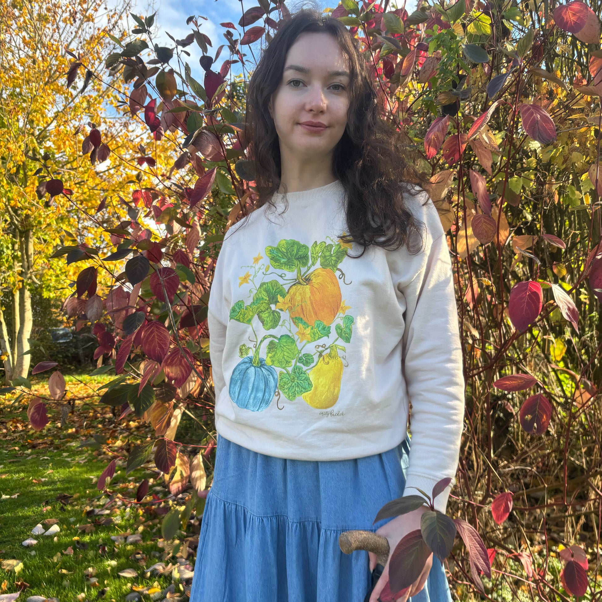 Gourd winter jumper with orange yellow and blue pumpkin illustrations and flowers. The colour of the jumper is light cream. The model wears it in front of a tree with red leaves. 