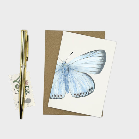 Featuring a light blue butterfly on an off-white card, layered over a brown kraft envelope. A gold pen and a small floral sticker are placed to the left on a light gray background.