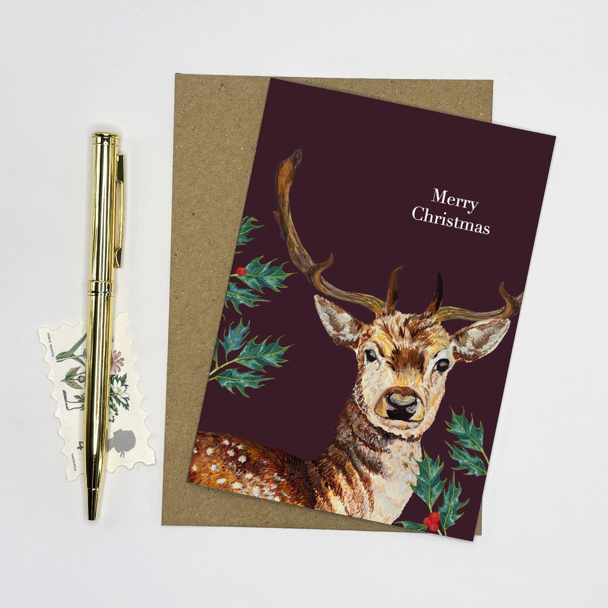A Christmas greeting card featuring an illustration of a deer with antlers and green holly leaves on a dark burgundy background, with 'Merry Christmas' printed in white. The card is shown with a brown envelope, a gold pen, and small decorative tags on a white background.