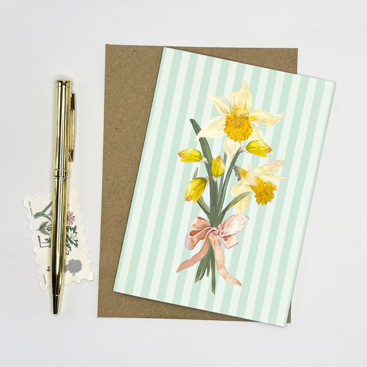 Daffodil Greeting card with wild daffodil flowers and a pink bow on a striped background, accompanied by a pen and stickers.
