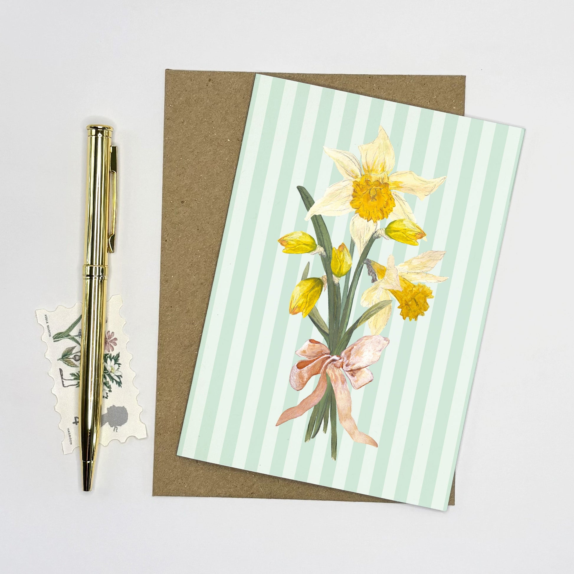 Daffodil Greeting card with wild daffodil flowers and a pink bow on a striped background, accompanied by a pen and stickers.