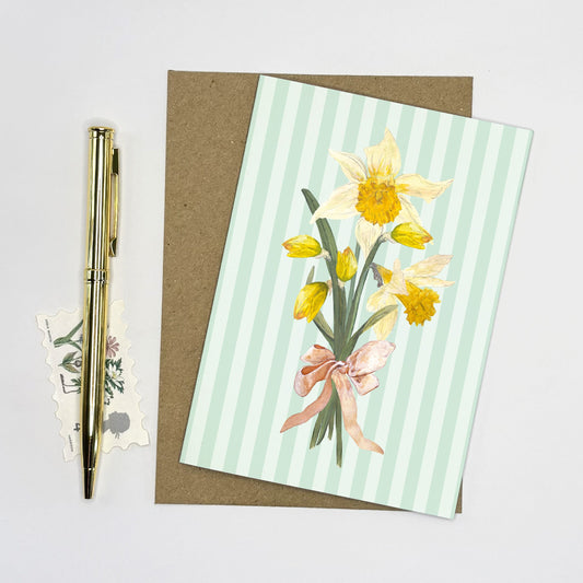 Daffodil Greeting card with wild daffodil flowers and a pink bow on a striped background, accompanied by a pen and stickers.