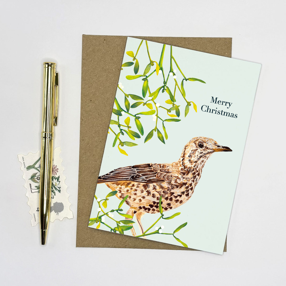 A Christmas greeting card featuring a watercolor bird perched among mistletoe with the text 'Merry Christmas,' shown with a brown envelope and a gold pen on a white background.