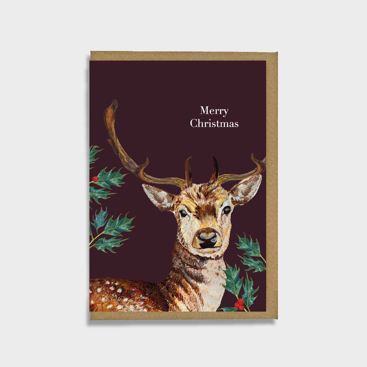 A Christmas greeting card featuring an illustration of a stag deer with antlers on a dark burgundy background, with green holly leaves and the text 'Merry Christmas' in white. The card has a brown kraft paper border and is shown against a light gray background.