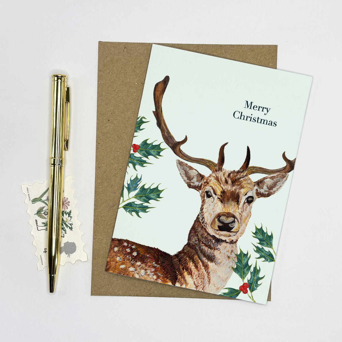 A Christmas greeting card featuring a watercolor illustration of a deer with antlers, surrounded by holly leaves and the text 'Merry Christmas,' shown with a brown envelope and a gold pen on a white background.