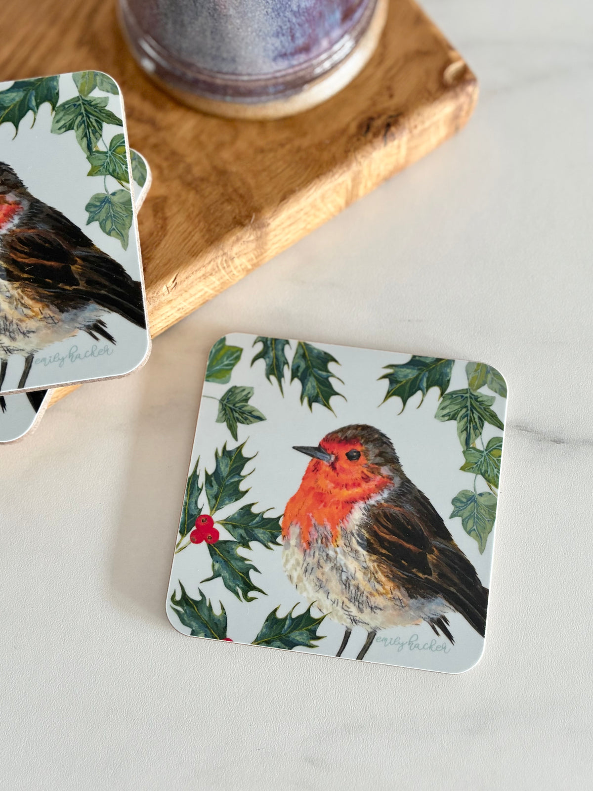 Robin melamine coaster with hand painted Robin, green Holly and ivy leaf illustrations on a light blue background. The coaster is photographed next to a wooden block and ceramic mug.