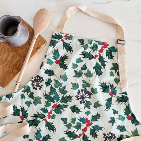 A white cotton apron with hand painted botanical holly and ivy illustrations, photographed next to a wooden block, wooden spoon and ceramic jug 