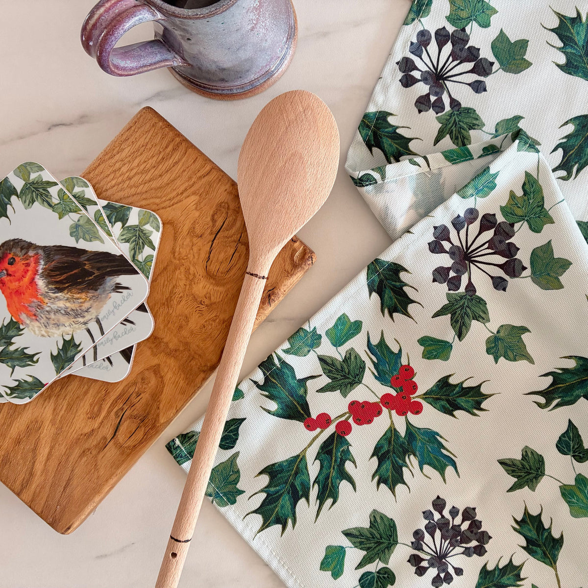 A styled flat-lay of a white tea towel printed with a green holly and red berry pattern, alongside a wooden spoon, a wooden cutting board, a bird-shaped coaster, and a ceramic mug on a light marble surface