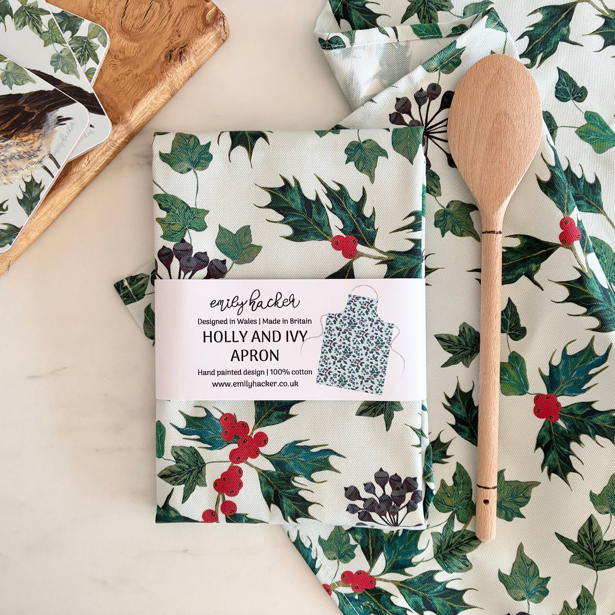 A folded cotton apron with a Holly and Ivy print is displayed on a light marble surface, accompanied by a wooden spoon and a matching printed coaster. The apron features a white base with green holly leaves and red berries, and has a white paper label wrapped around the middle.