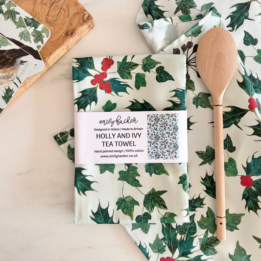 A folded tea towel with a green holly and red berry pattern is wrapped in a paper band labeled 'Emily hacker' and 'HOLLY AND IVY TEA TOWEL,' placed on a light marble surface with a wooden spoon and matching printed napkins nearby.