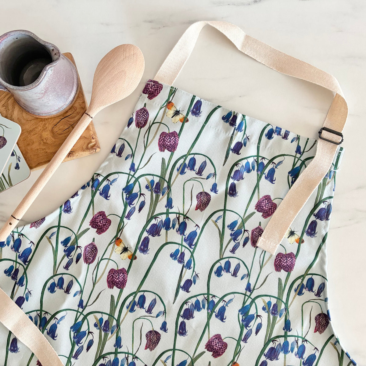 A floral kitchen apron with a bluebell and snake head fritillary floral print, styled in a flat lay with a wooden spoon, ceramic mug, and matching coasters on a light marble surface.