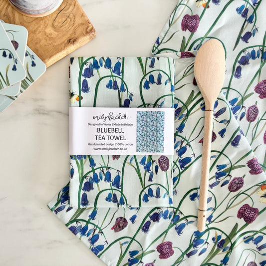 A cotton tea towel with hand painted bluebell and snake head fritillary design on a light blue background. It is photographed next to matching coasters, a wooden spoon and a wooden chopping board.