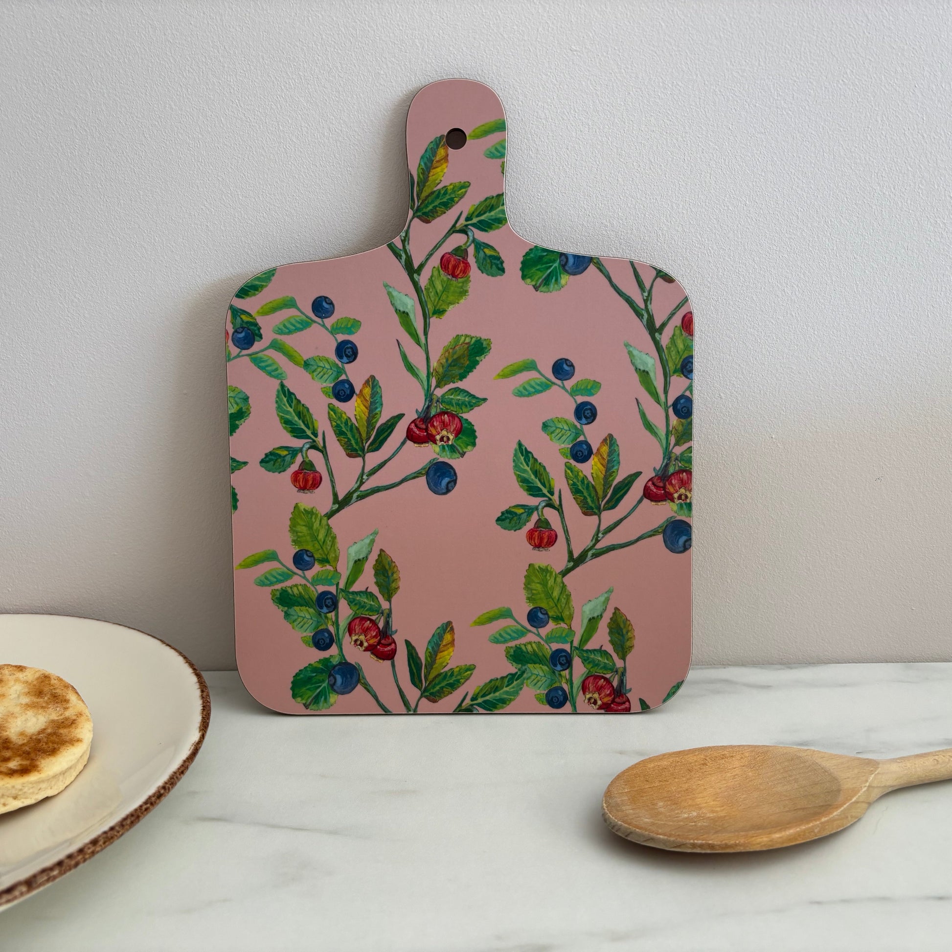 Pink cutting board with floral and berry design on a white surface with wooden spoon and plate