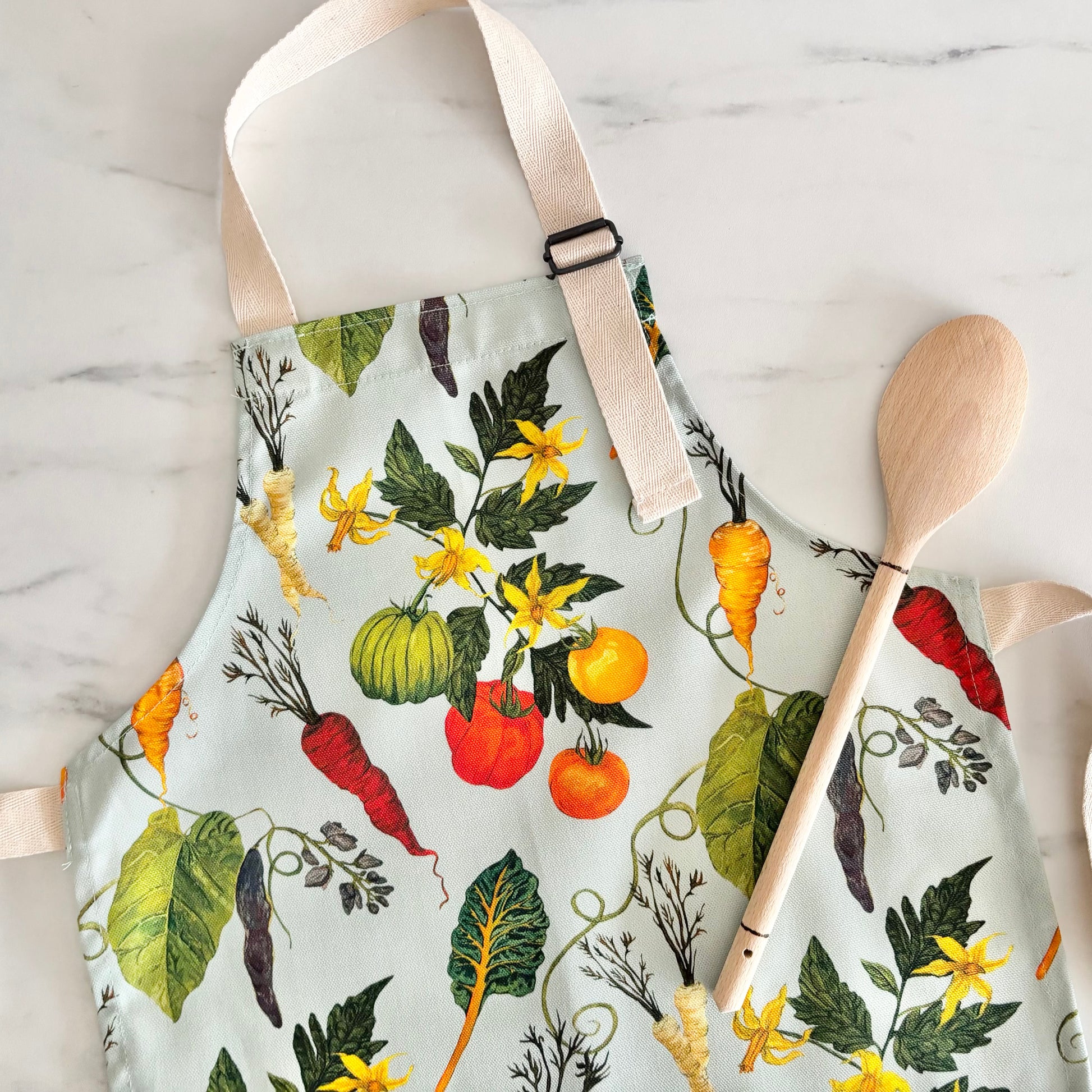 Kids cotton apron with illustrative vegetable pattern and wooden spoon on marble surface