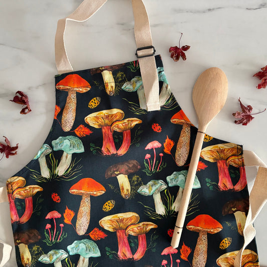 Hand designed kids apron with mushroom pattern on a marble surface with a wooden spoon and leaves.