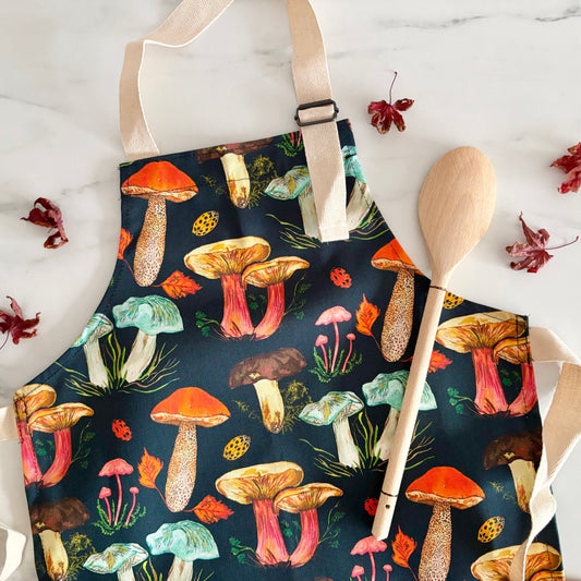 Hand designed cotton kids apron with mushroom pattern on a marble surface with a wooden spoon and leaves.