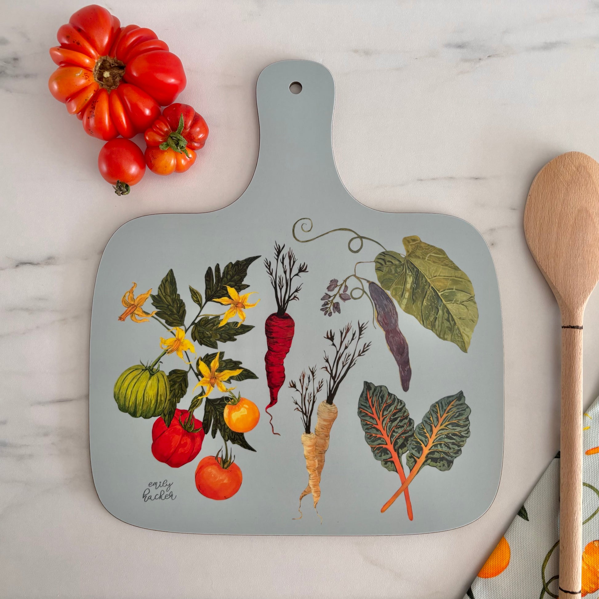 Light blue cutting board with heritage and heirloom vegetable illustrations on a marble surface with tomatoes and a wooden spoon