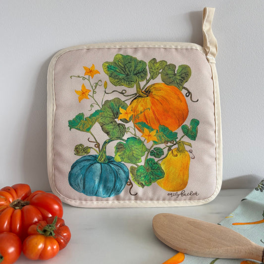 Potholder with pumpkin design on a white surface with tomatoes and a wooden spatula.