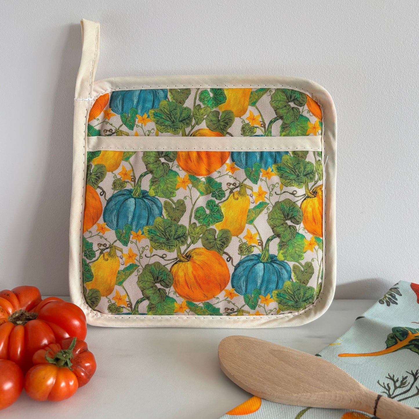 Potholder with pumpkin pattern on a white surface with tomatoes and a wooden spoon.