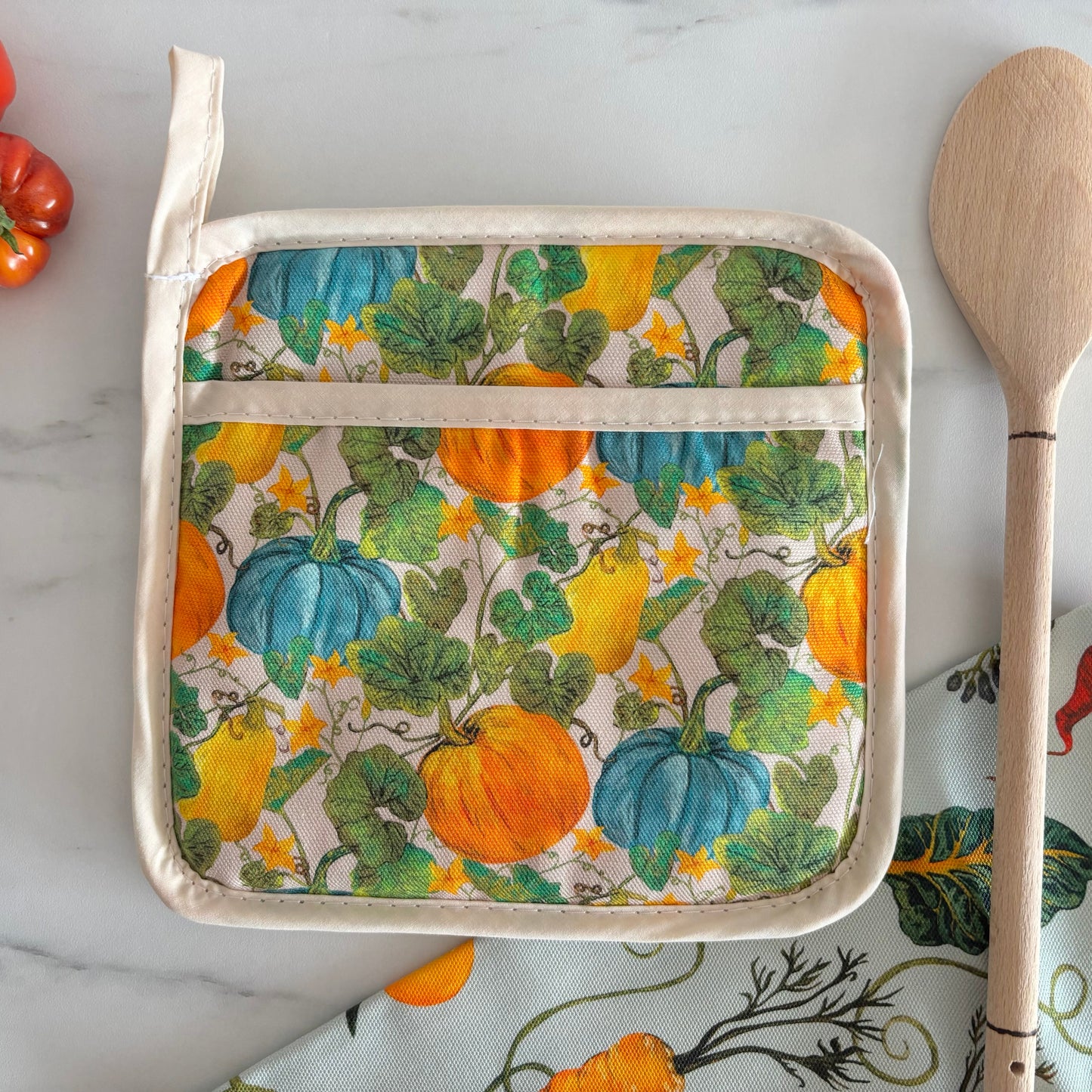 Potholder with pumpkin pattern on a marble surface with a wooden spoon and tomatoes.
