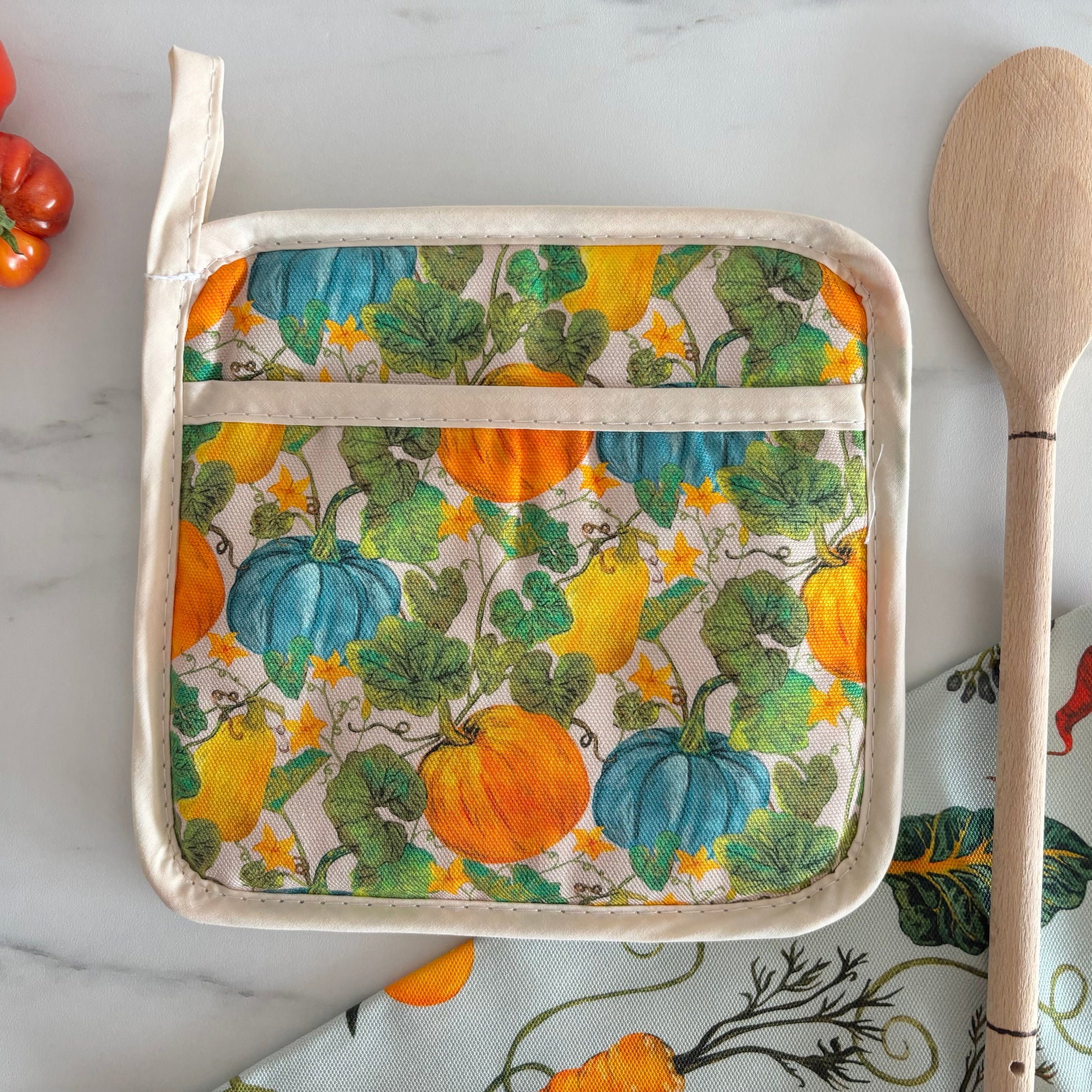 Potholder with pumpkin pattern on a marble surface with a wooden spoon and tomatoes.