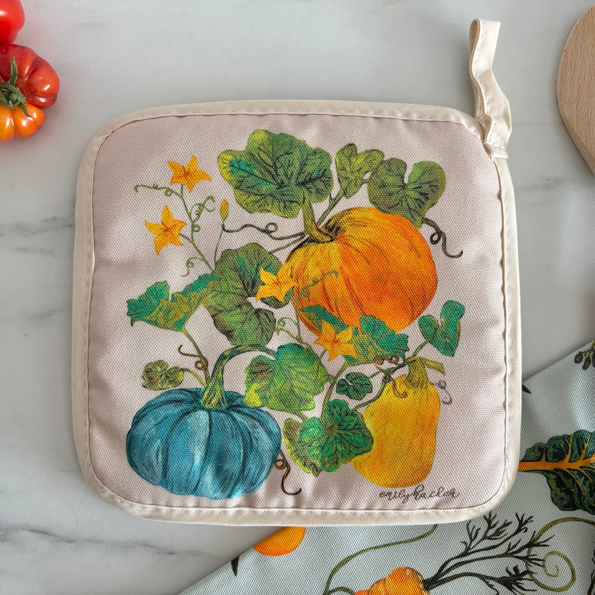 Potholder with pumpkin design on a marble surface with tomatoes and a wooden spoon.