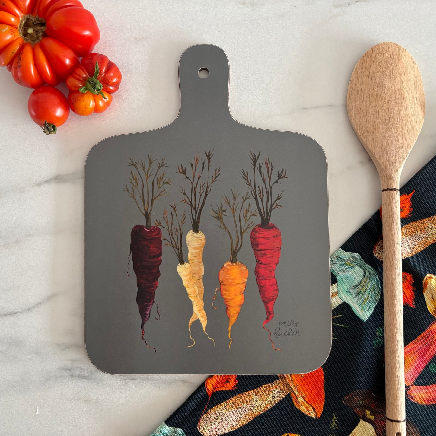 Grey cutting board with hand painted heritage carrot design photographed next to wooden spoon, and tomatoes on a marble surface