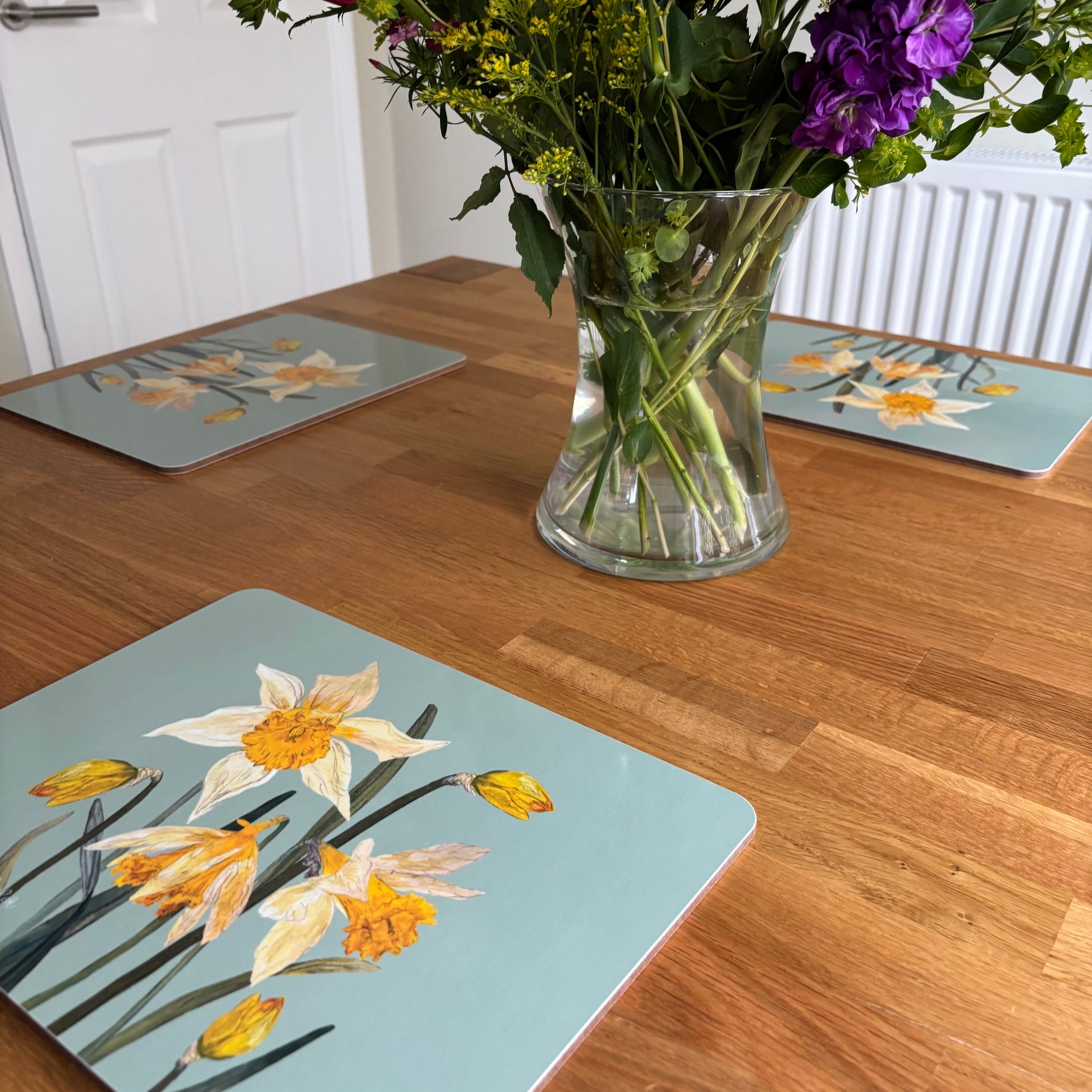 Table with floral wild daffodil duckegg blue placemats and a vase of flowers on a wooden table.