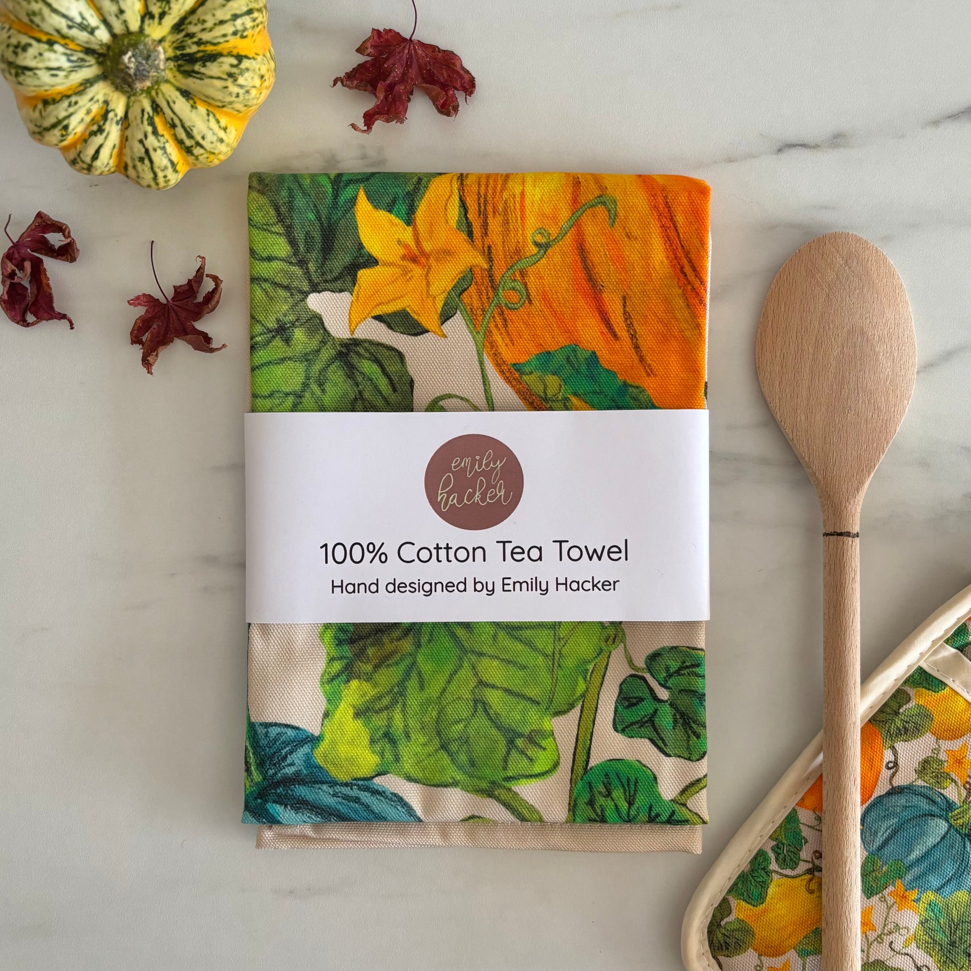 100% cotton tea towel with pumpkin and squash design in its bellyband packaging it is next to a gourd, autumn leaves and wooden spoon on marble surface