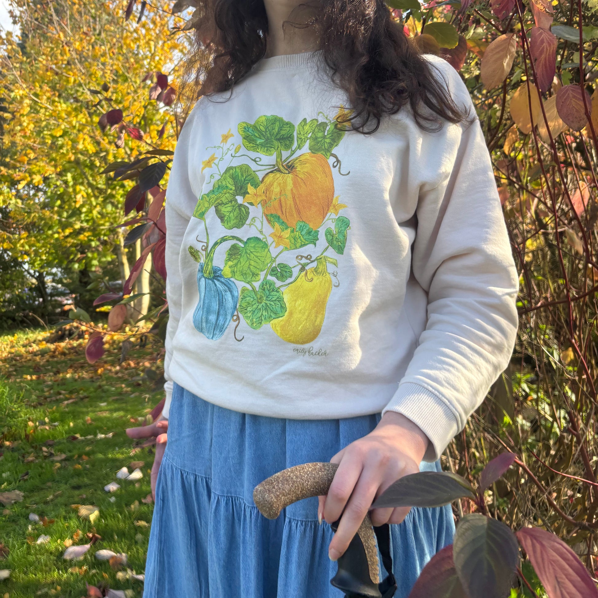 Cream jumper with multicolour autumnal pumpkin squash and gourd illustrations. The model wears the jumper in front of a tree with brown winter leaves.