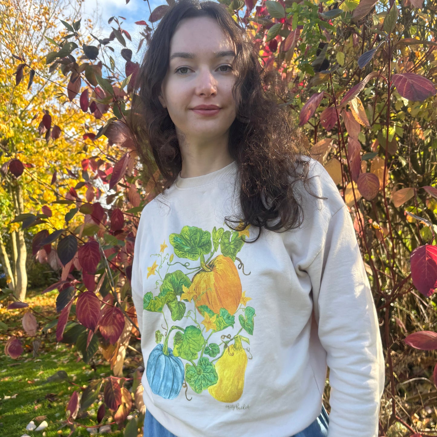 Person wearing a white sweater with a colorful pumpkin design, standing in front of a tree with autumn leaves.