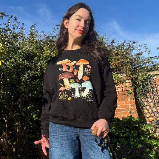 Organic cotton Black jumper with woodland mushroom design. A Women standing outdoors with a scenic background of trees and clear sky is wearing the jumper.