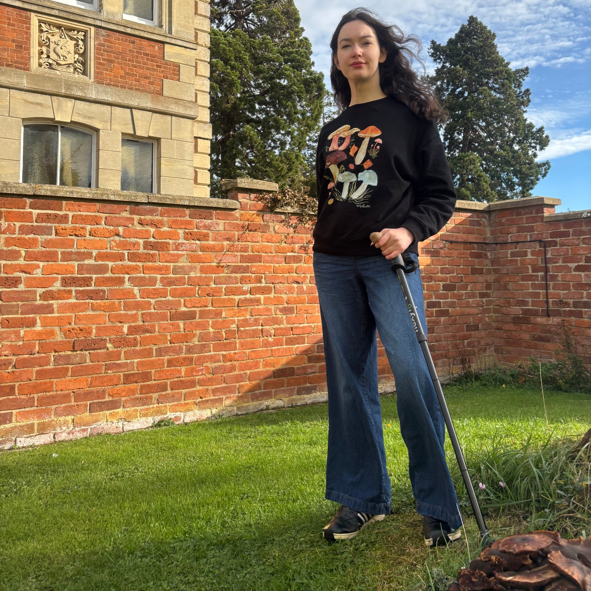 Black multicoloured fungi sweatshirt. The Person is wearing the organic cotton jumper standing outdoors with a building and greenery in the background