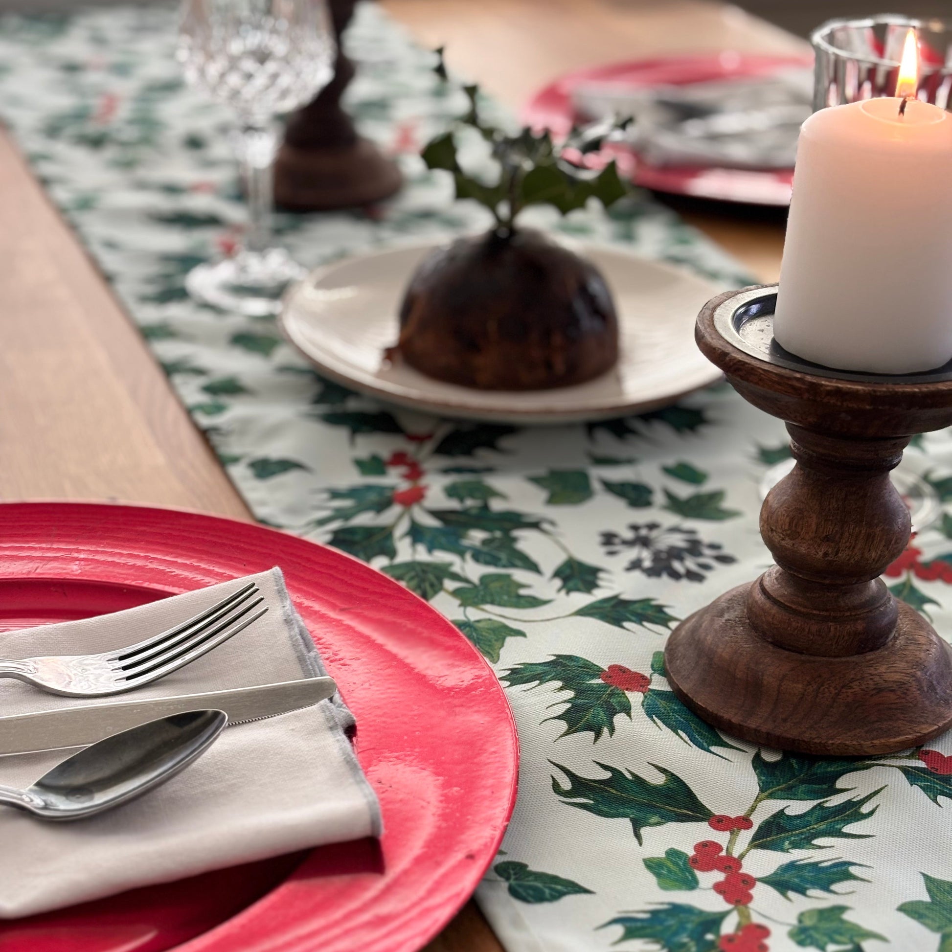 Christmas table setting with red plates, holly and ivy leaf tablecloth, and wooden candle holder.