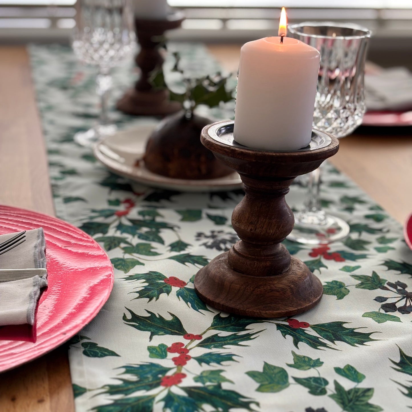 Christmas Table setting with a decorative tablecloth featuring ivy holly leaves and berries, a candle in a wooden holder, and red plates.