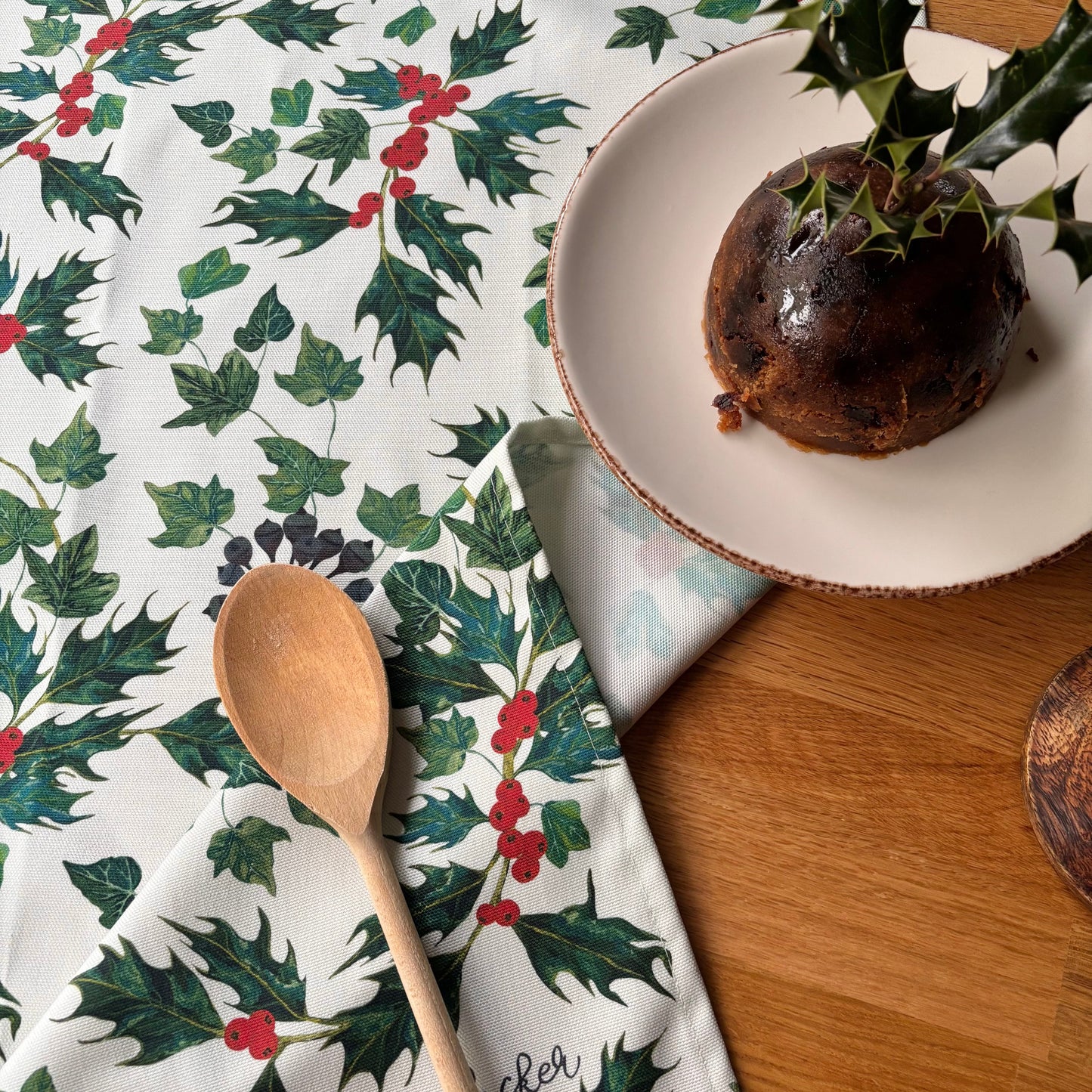Christmas Holly and ivy tea towel next to Christmas pudding and a wooden spoon