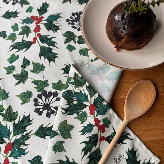 Holly leaf patterned tea towel with a dish, wooden spoon, and napkin on a wooden surface.