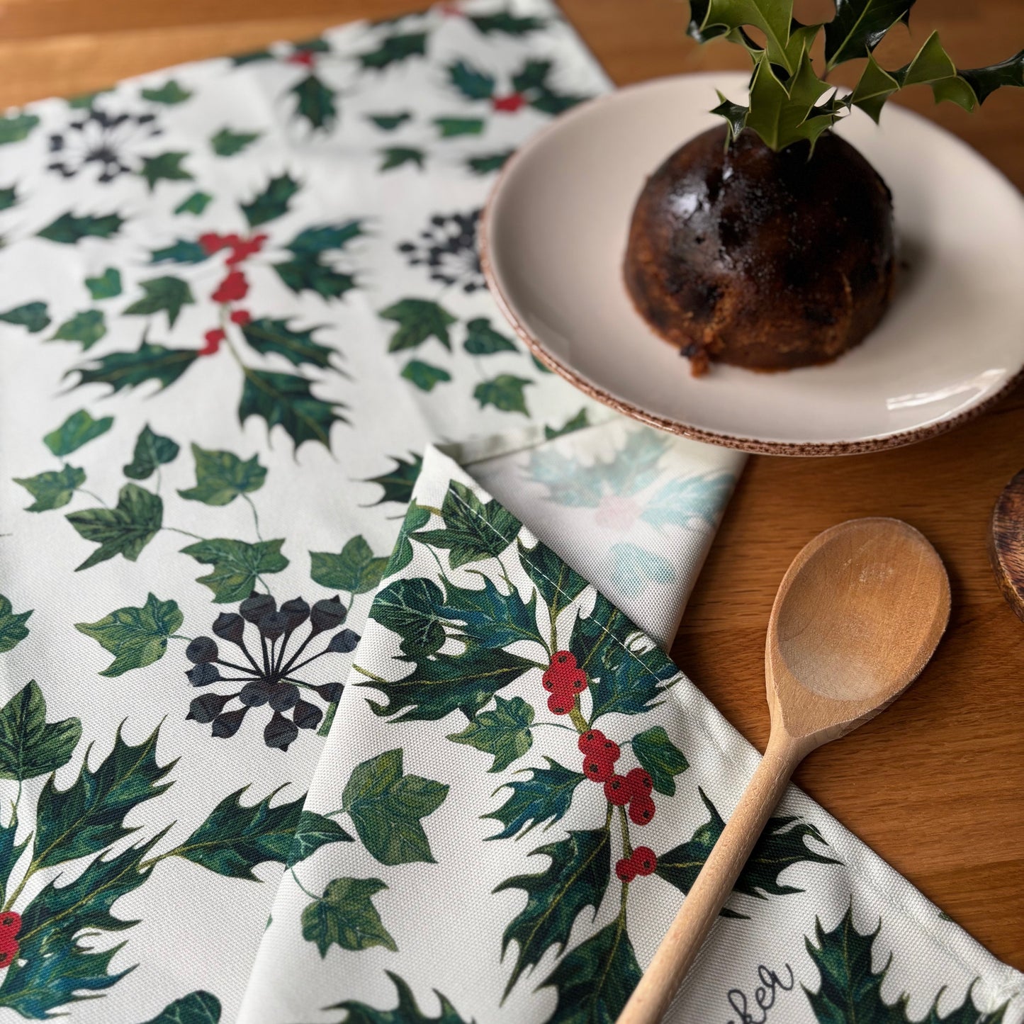 Holly and ivy leaf patterned tea towel  on a wooden table with a wooden spoon and Christmas pudding