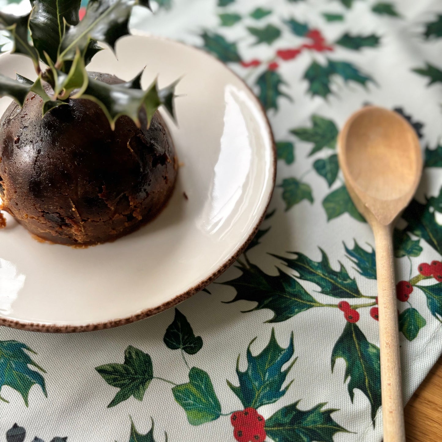 Christmas pudding on a white plate with a wooden spoon on a holly leaf patterned tea towel.