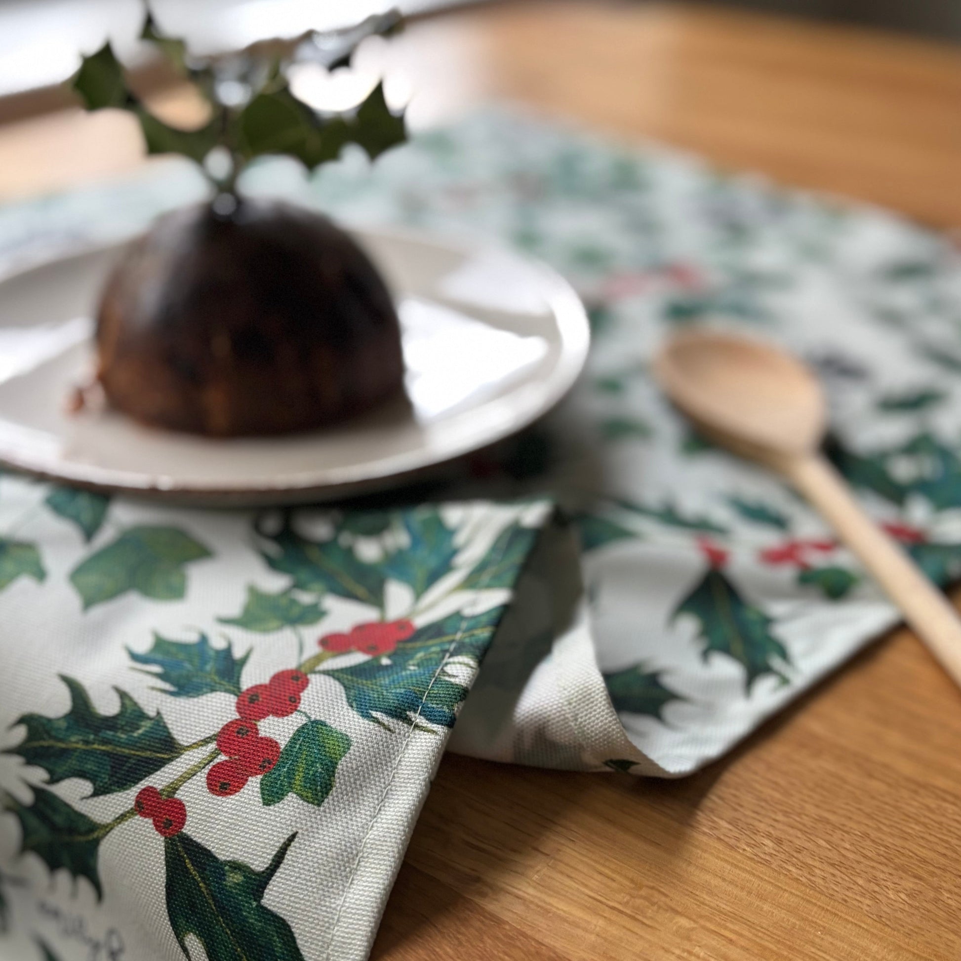 Christmas-themed table setting with a plum pudding on a plate, surrounded by holly berries and ivy leaves tea towel and wooden spoon