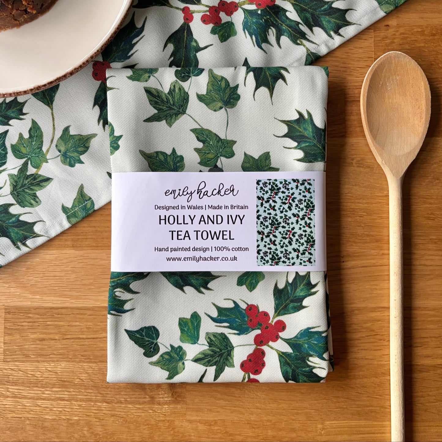 Tea towel with holly and ivy design on a wooden surface with a wooden spoon and plate.