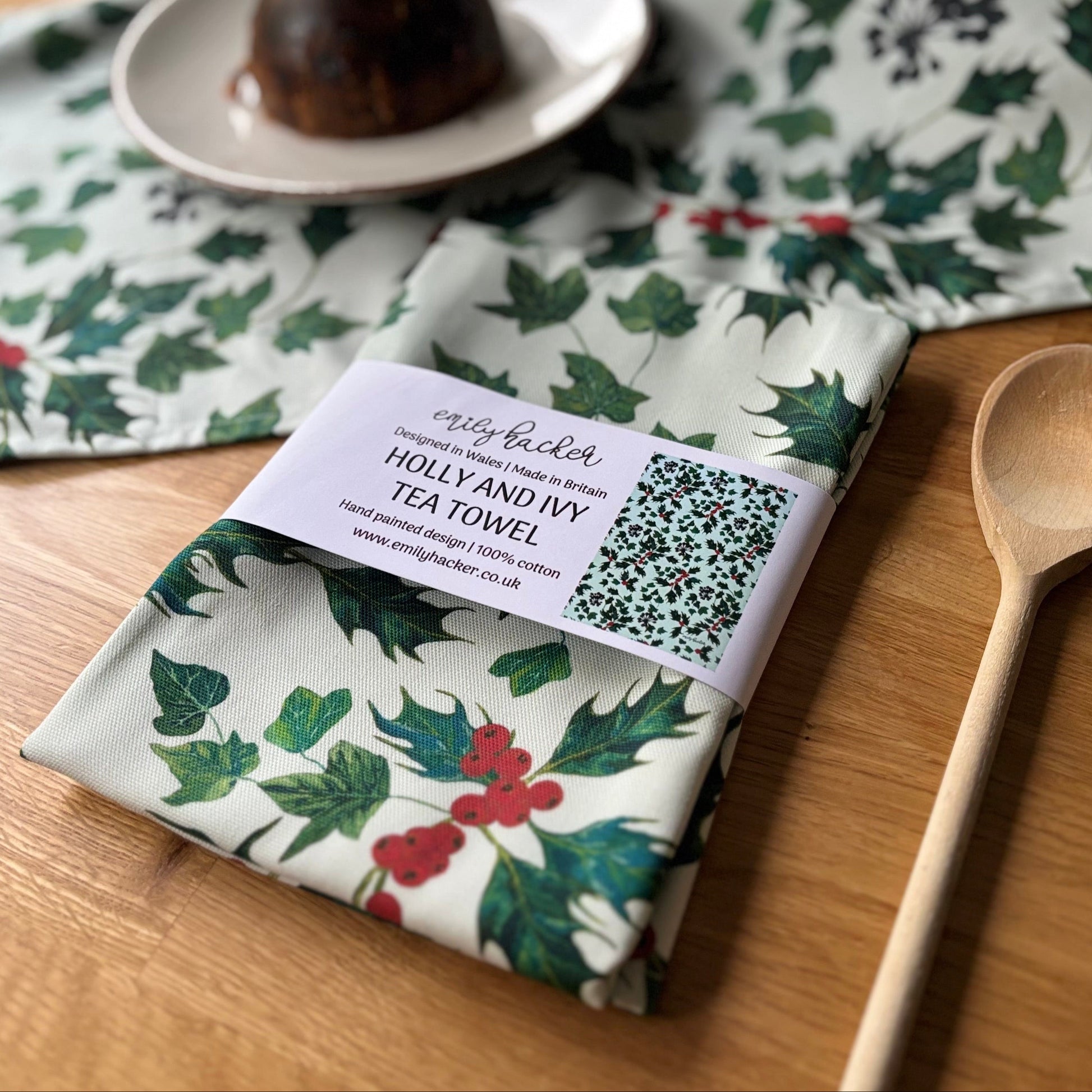 Tea towel with holly and ivy design on a wooden table with a plum dessert and wooden spoon.