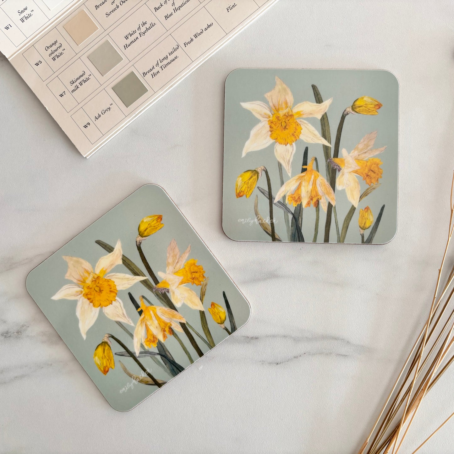 Two floral coasters with daffodils on a marble surface with dried plants.