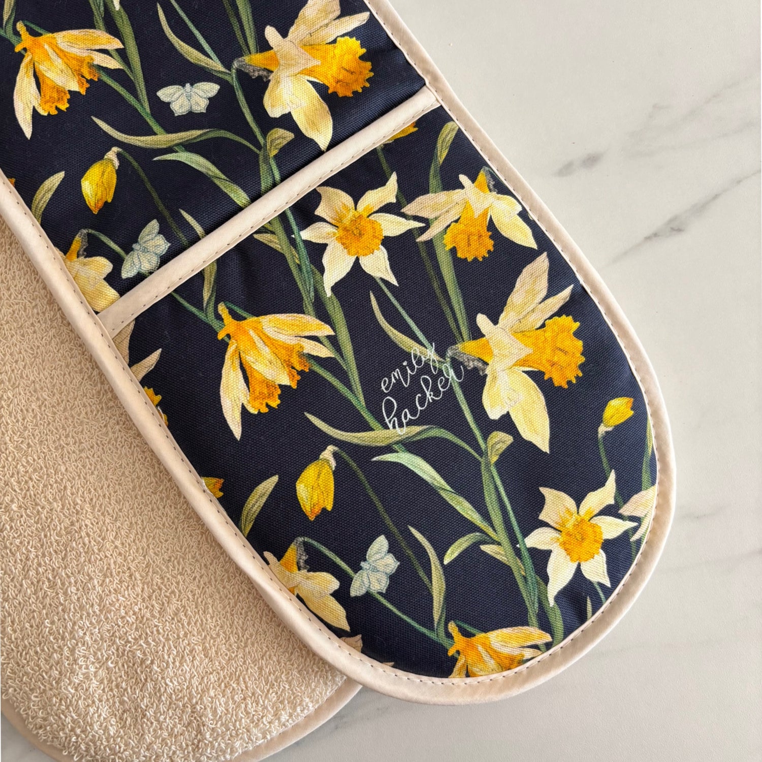 A double oven glove featuring hand painted illustrations of wild daffodils on a black background, with a beige towelled reverse.